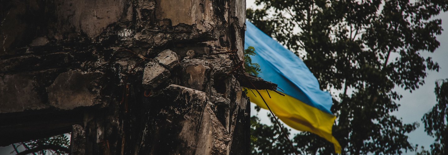 A Ukrainian flag waves in the background of war-torn ruins, captured with an artistic blur effect. The image symbolizes resilience amidst destruction during the war in Ukraine