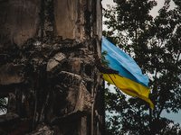 A Ukrainian flag waves in the background of war-torn ruins, captured with an artistic blur effect. The image symbolizes resilience amidst destruction during the war in Ukraine