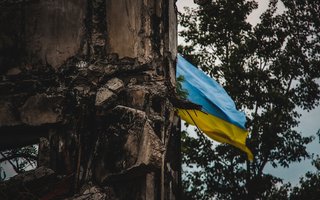 A Ukrainian flag waves in the background of war-torn ruins, captured with an artistic blur effect. The image symbolizes resilience amidst destruction during the war in Ukraine