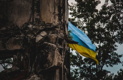 A Ukrainian flag waves in the background of war-torn ruins, captured with an artistic blur effect. The image symbolizes resilience amidst destruction during the war in Ukraine