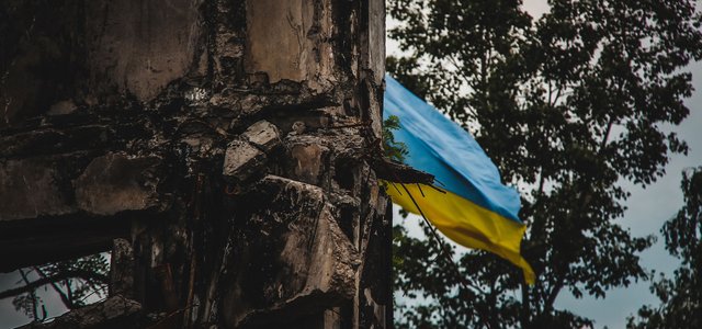 A Ukrainian flag waves in the background of war-torn ruins, captured with an artistic blur effect. The image symbolizes resilience amidst destruction during the war in Ukraine
